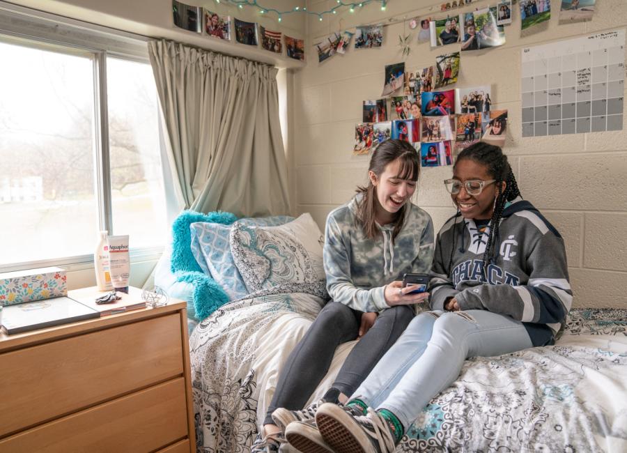 two students sitting on a bed near a window in a dorm room